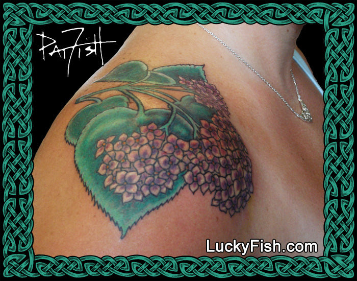 Tattoo of purple hydrangea flowers and green leaves by Pat Fish on a person's shoulder with LuckyFish branding.