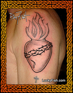 A black and gray tattoo of the Sacred Heart of Jesus with flames, applied to a person's upper arm. The image features a decorative Celtic-style border and includes a url 'LuckyFish.com'.
