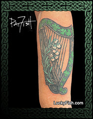 Tattoo of a green harp with flowers on a person's arm with Celtic knotwork and 'LuckyFish.com' branding