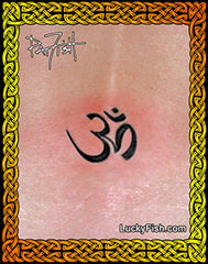A black Om symbol tattoo design centered on a pink skin background, surrounded by a decorative gold and orange Celtic-style border. A stylized signature appears in the top left, and 'LuckyFish.