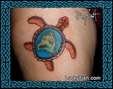 A close-up of a tattoo on skin showing a sea turtle with a blue ocean and Hawaiian island design inside its shell. The photo is framed by a blue Celtic-style border, with the text LuckyFish.