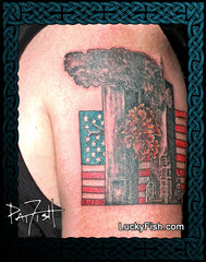 A close-up of a shoulder with a large tattoo depicting two smoking towers, an American flag, a fireball, and city buildings. The photo includes a decorative dark blue Celtic knot border, a tattoo artist signature reading Pat Fish, and the website LuckyFish.