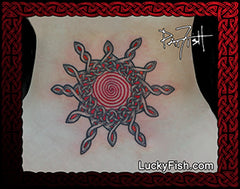 A circular Celtic knot tattoo design on skin, featuring a red spiral center with dark gray and red interwoven geometric patterns. The tattoo is displayed on a lower back with a red Celtic border frame, and a watermark 'LuckyFish.