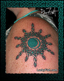 A close-up of a shoulder with a black Celtic knot circular sunburst tattoo featuring a teal-green center, surrounded by a decorative dark green border and the text LuckyFish.com.